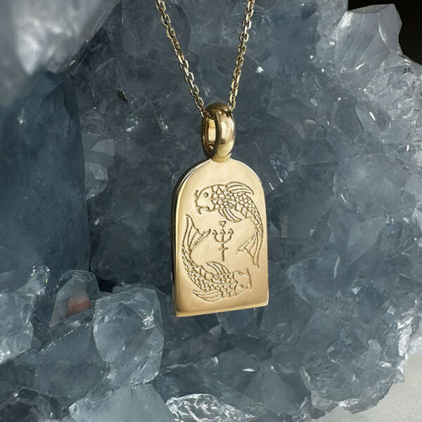 yellow gold pendant hanging in front of blue crystal. Pendant is embossed with two koi fish, juju gems original Pisces design.